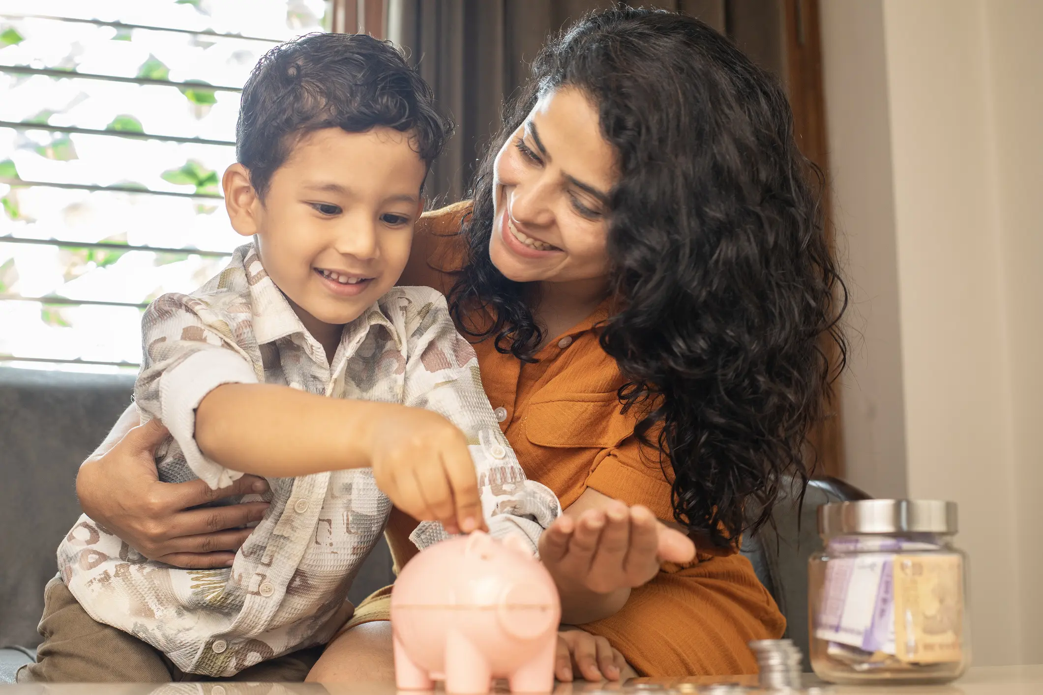 Smiling young mother inserting coins in piggy bank while sitting on sofa in living room