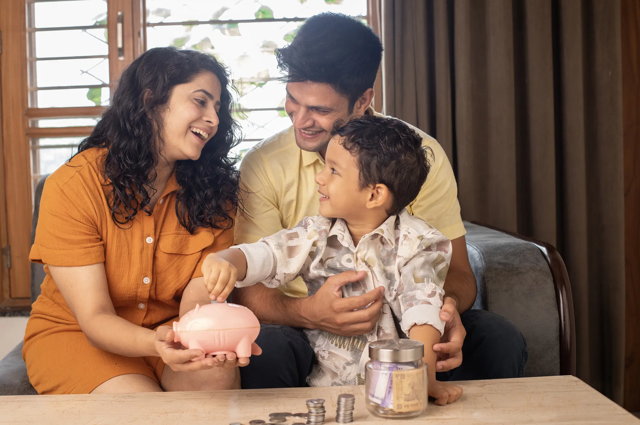 Cheerful parents looking at smiling son inserting coins in piggy bank and saving money in living room at home